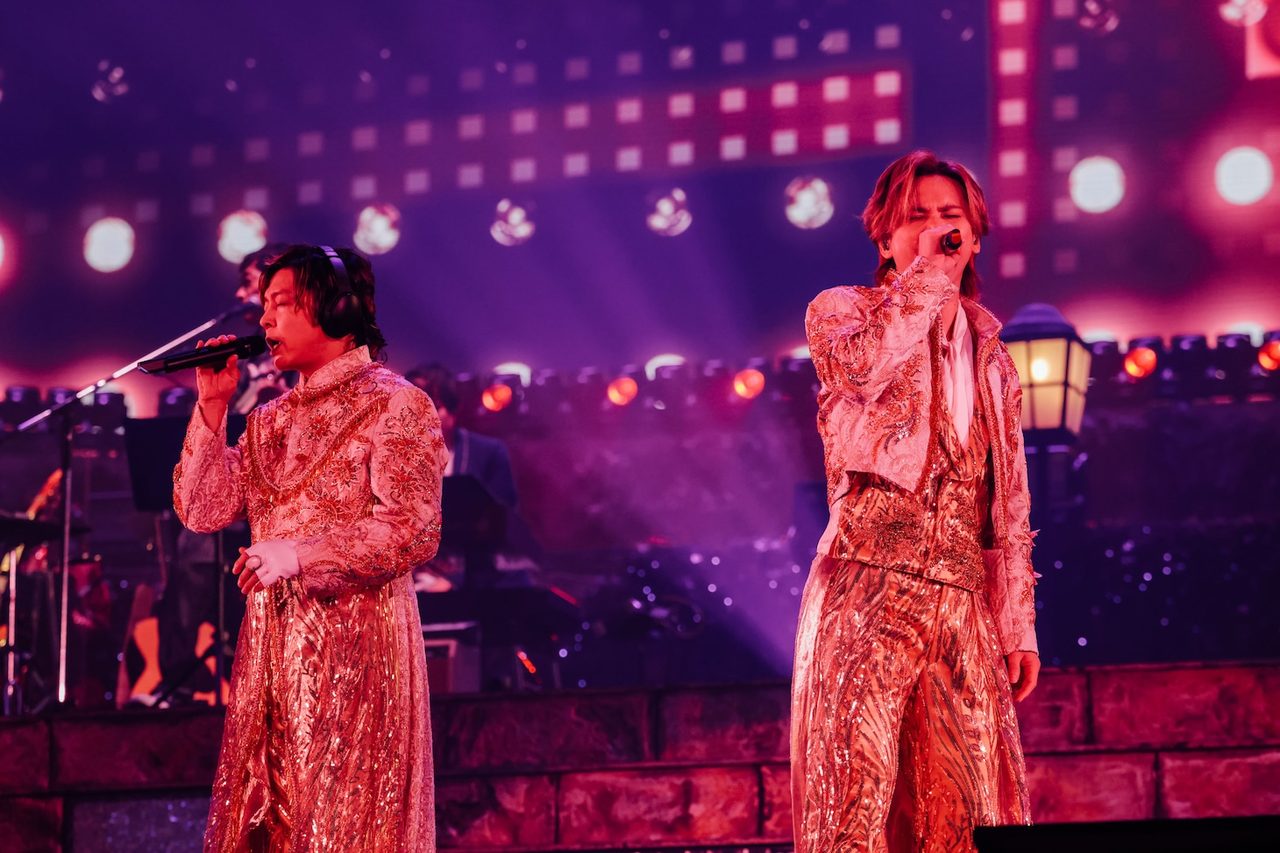 DOMOTO堂本光一と堂本剛がゴールドの衣装で歌唱するステージ、背景に赤とピンクの照明演出