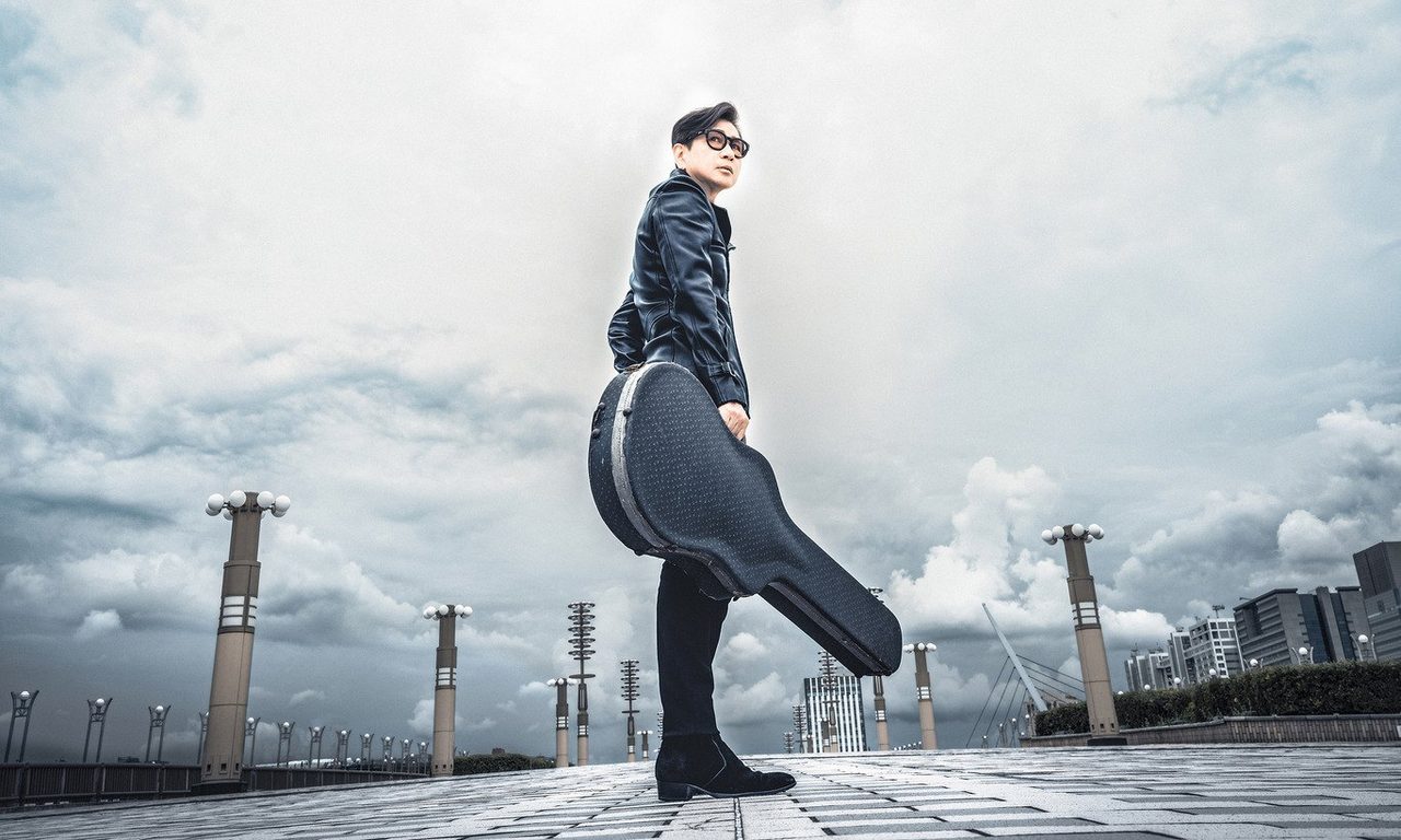黒いレザージャケット姿でギターケースを持つ藤井フミヤのアーティスト写真
