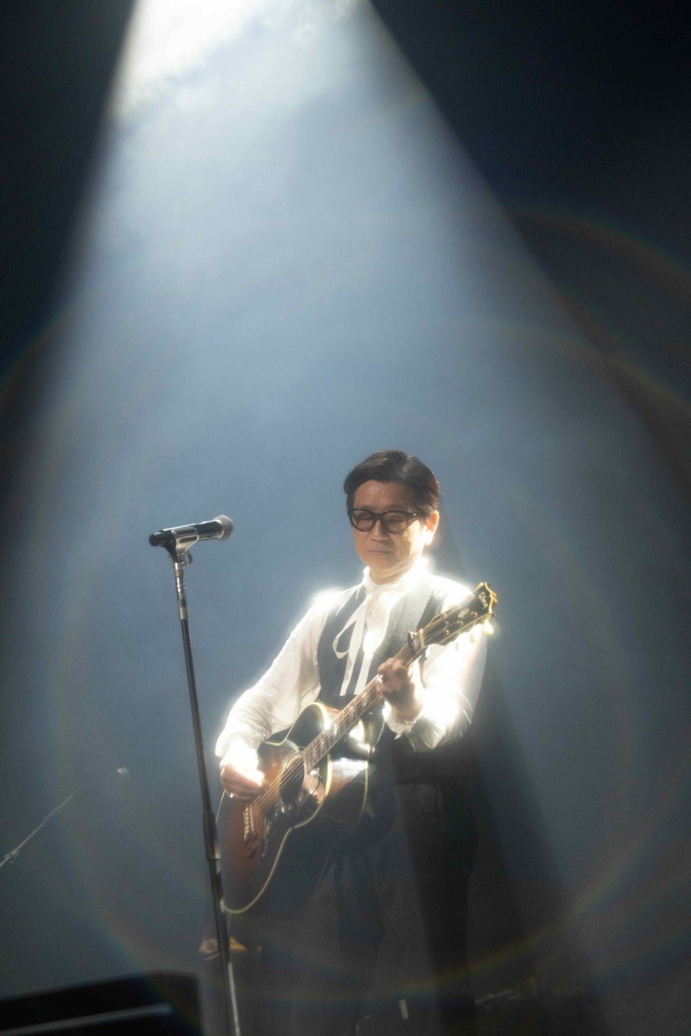 スポットライトの中でアコースティックギターを弾く藤井フミヤのライブ写真