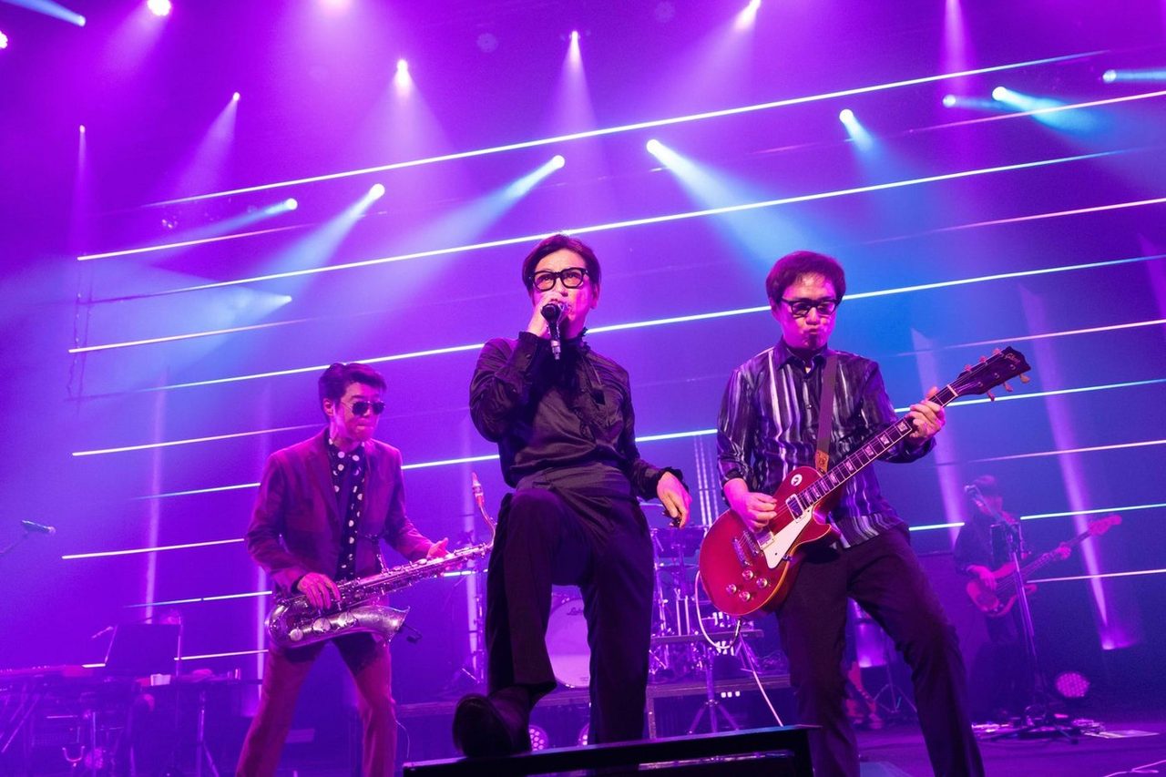 ステージで歌う藤井フミヤと、サックスの藤井尚之、ギター奏者が並ぶ紫の照明のライブ写真