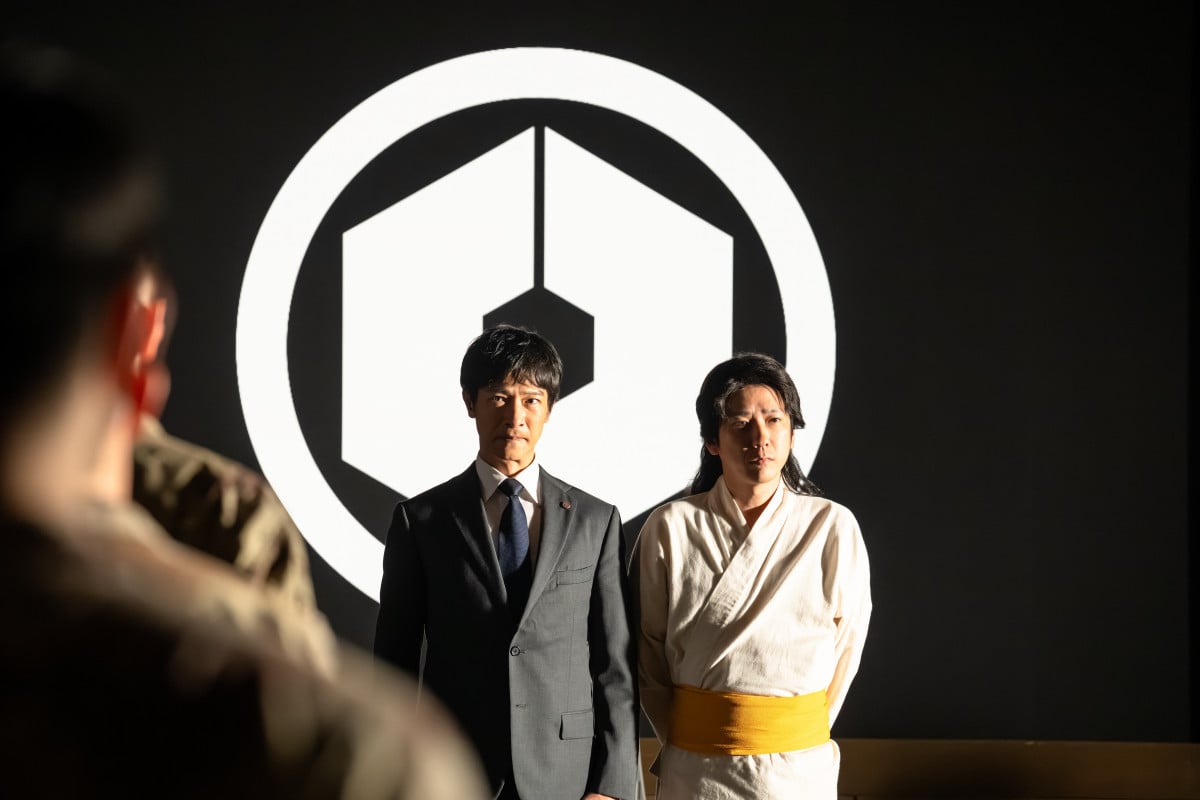 VIVANT第2シリーズ 六角形の紋章の前に並んで立つスーツの男性と白い衣装の女性