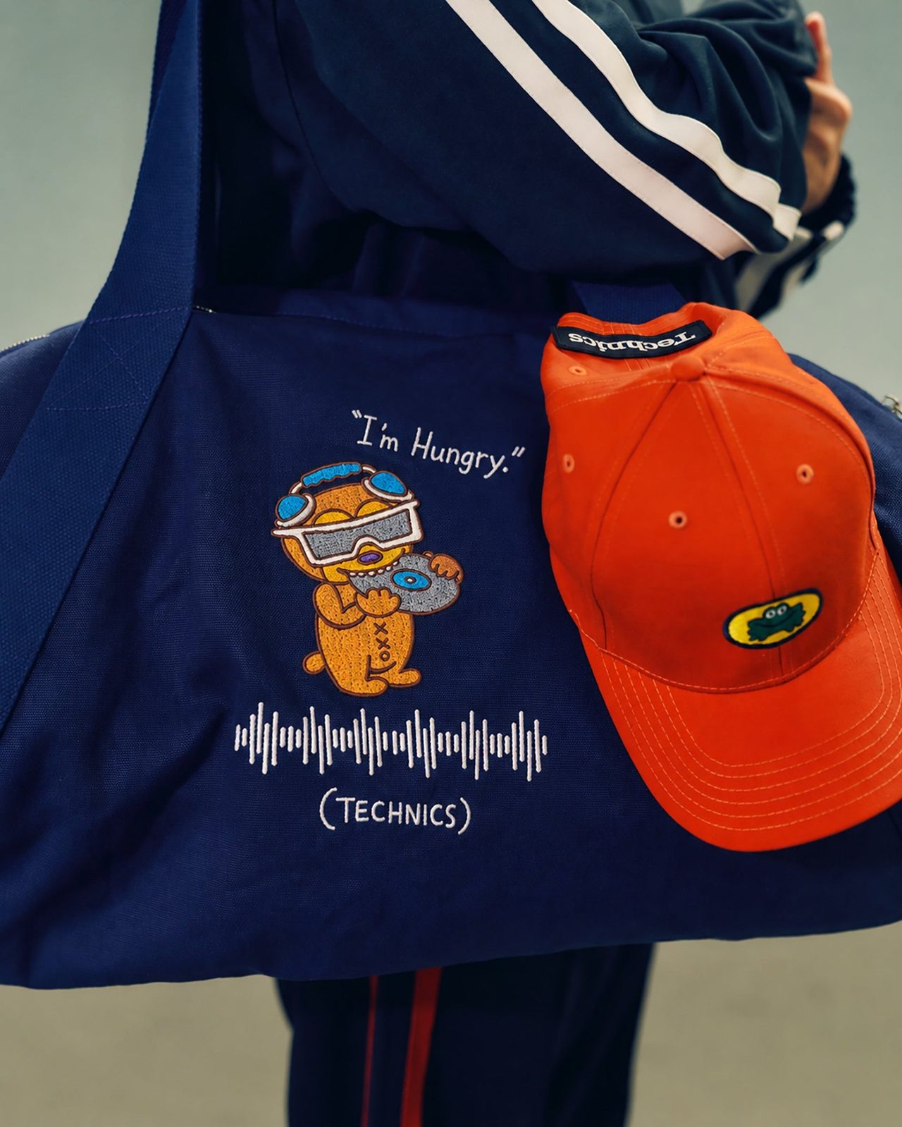 ネイビーのバッグにキャラクターのイラストと「I'm Hungry.」の文字、オレンジのキャップ
