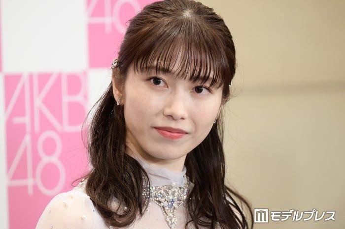 AKB48のロゴを背景に微笑む横山由依さんのバストアップ写真